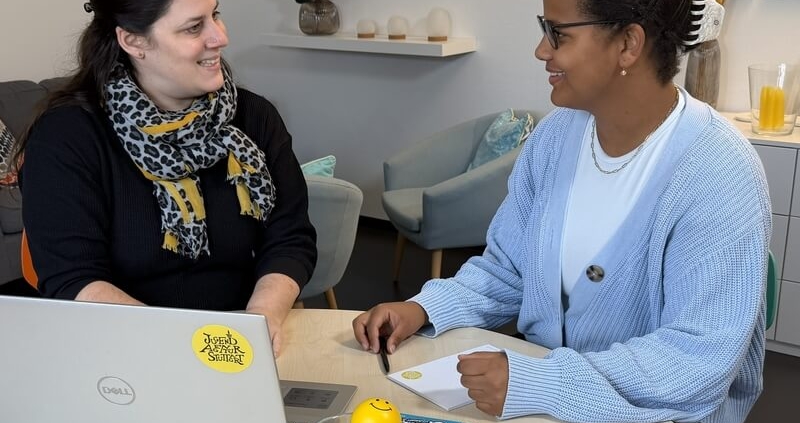 Zwei Frauen sitzen an einem runden Tisch mit Laptop, Notizblock, Flyern und einem gelben Smiley-Ball, im Hintergrund Sessel und Wandregal.