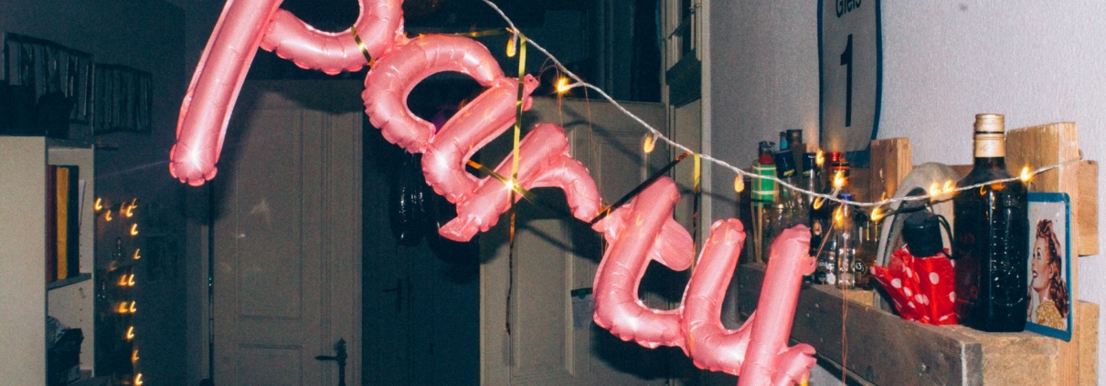 Luftballon-Buchstaben mit Lichterkette formen das Wort 'party' in einem Flur mit Regalen und Flaschen.