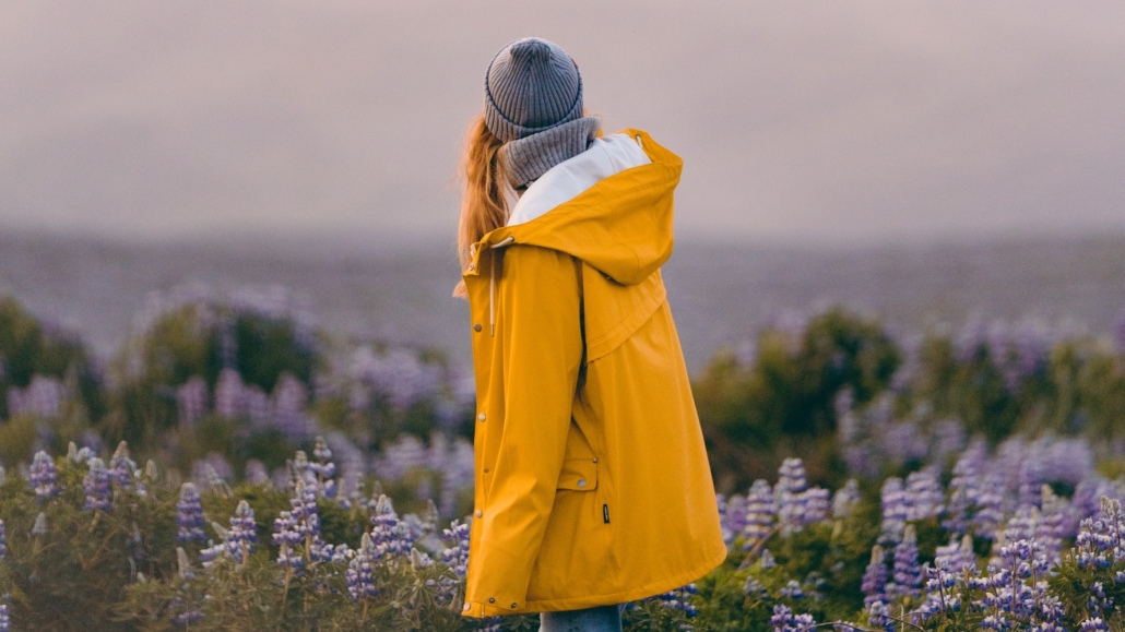 Eine junge Frau im gelben Regenmantel schaut auf ein Blumenfeld
