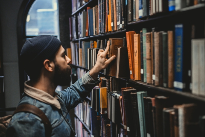 Junger Mann mit Mütze steht an einem Bücherregal und nimmt sich ein altes Buch